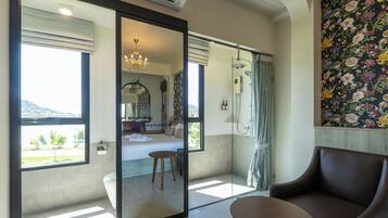 Una habitación de hotel moderna con un gran ventanal corredero que da a un balcón, una cama, zona de ducha y un área de descanso con una mesa.