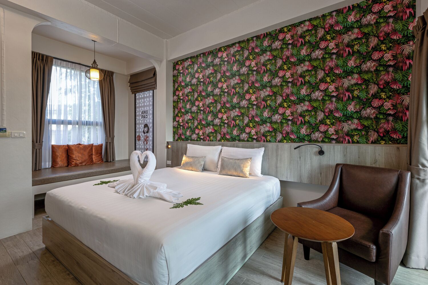 Habitación de hotel con una cama grande, cabecera de madera, un sillón de cuero marrón, una mesita de madera y un papel tapiz con motivos de hojas tropicales.