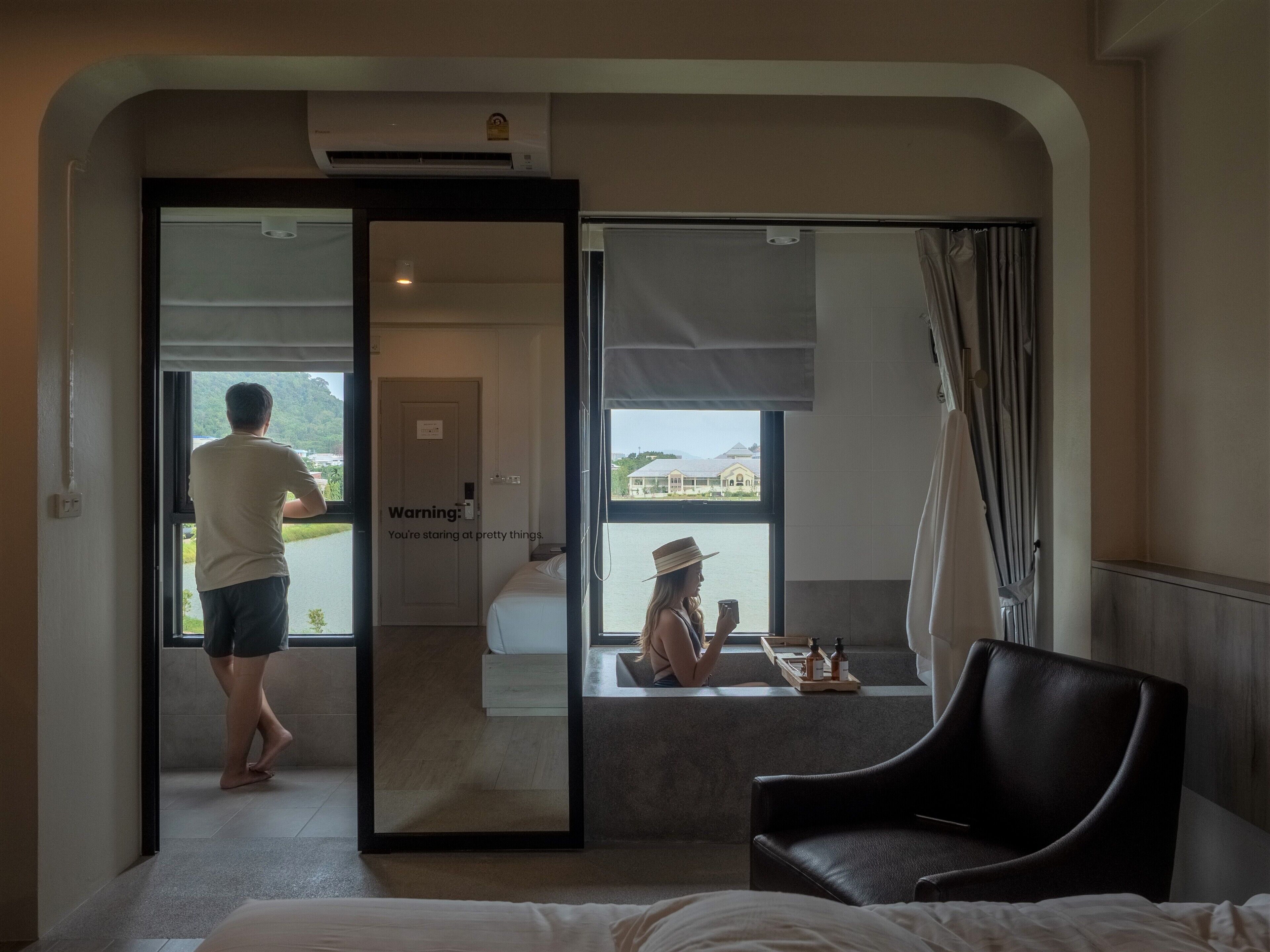 A hotel room with a bed, a chair, a window with a view of buildings, and a door leading to another room.