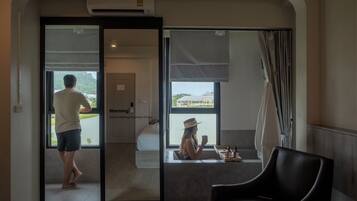 A hotel room with a bed, a chair, a window with a view of buildings, and a door leading to another room.