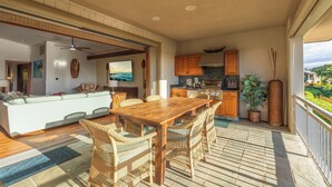 A spacious living area with a wooden dining table and wicker chairs, a kitchen with wooden cabinets, and a balcony with a view of the landscape. - Ohana Paradise@ Mauna Lani Villages, private beach club access (Waimea)
