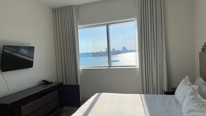 A hotel room with a bed, a TV mounted on the wall, a desk with a chair, and a window with a view of a city skyline and water. - Lovely waterfront condo (Hollywood)
