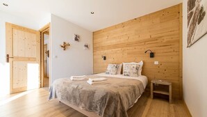 Un dormitorio con cabecera de madera, una cama con manta gris, una mesita de noche y una puerta que da a otra habitación.