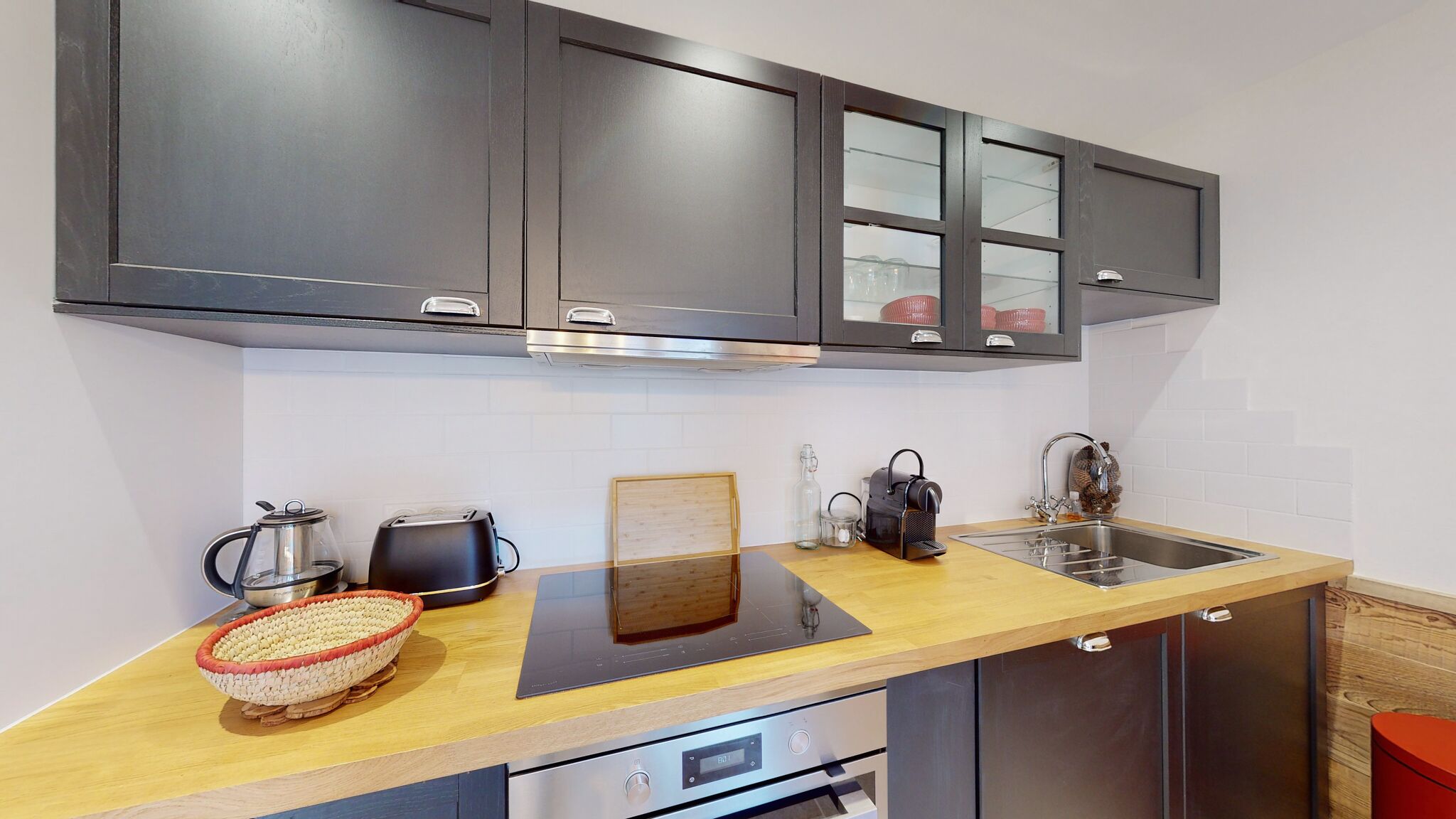 Una cocina moderna con armarios oscuros, encimera de madera, horno empotrado, pileta y un taburete rojo.
