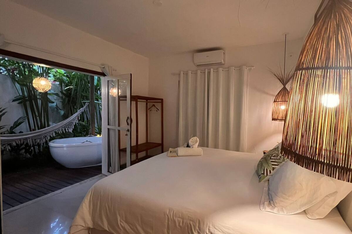 Quarto com uma cama grande, mesa de cabeceira, luminária pendente e vista para um banheiro com banheira.