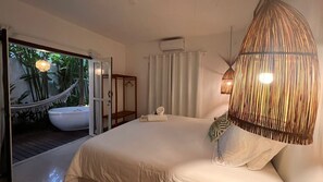 A bedroom with a large bed, a bedside table, a hanging light fixture, and a view of a bathroom with a bathtub. - Pousada Bem te vi trancoso (Porto Seguro)