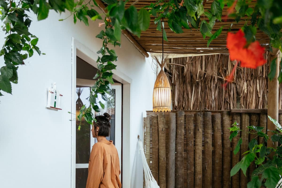 Um deck de madeira com uma rede, uma pessoa parada numa porta e um muro de bambu.