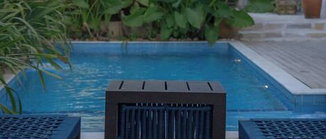 Una terraza de madera con piscina, rodeada de exuberante vegetación y un edificio con detalles azules.