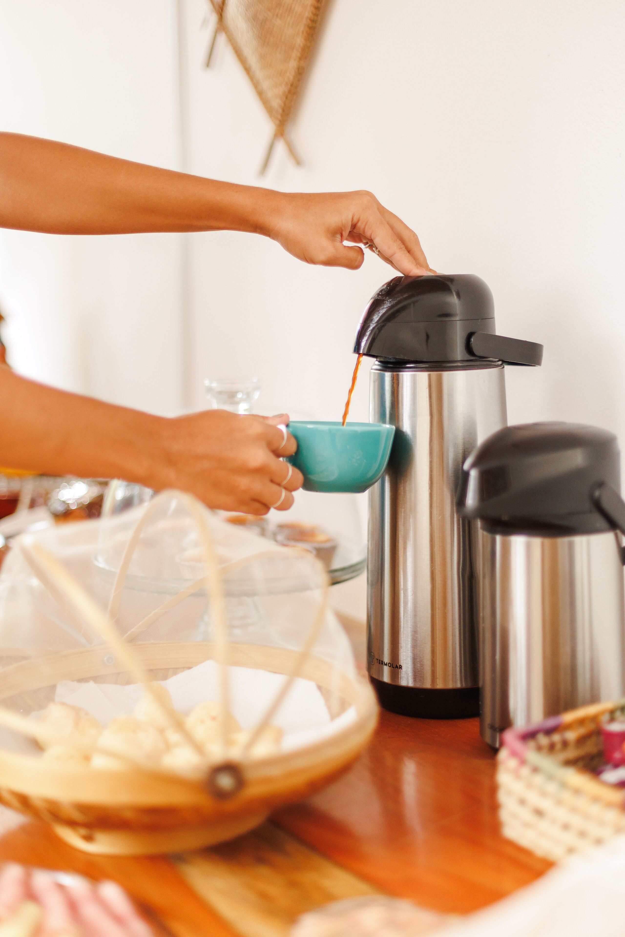 Una persona sirve café desde una cafetera de acero inoxidable en una taza azul turquesa.