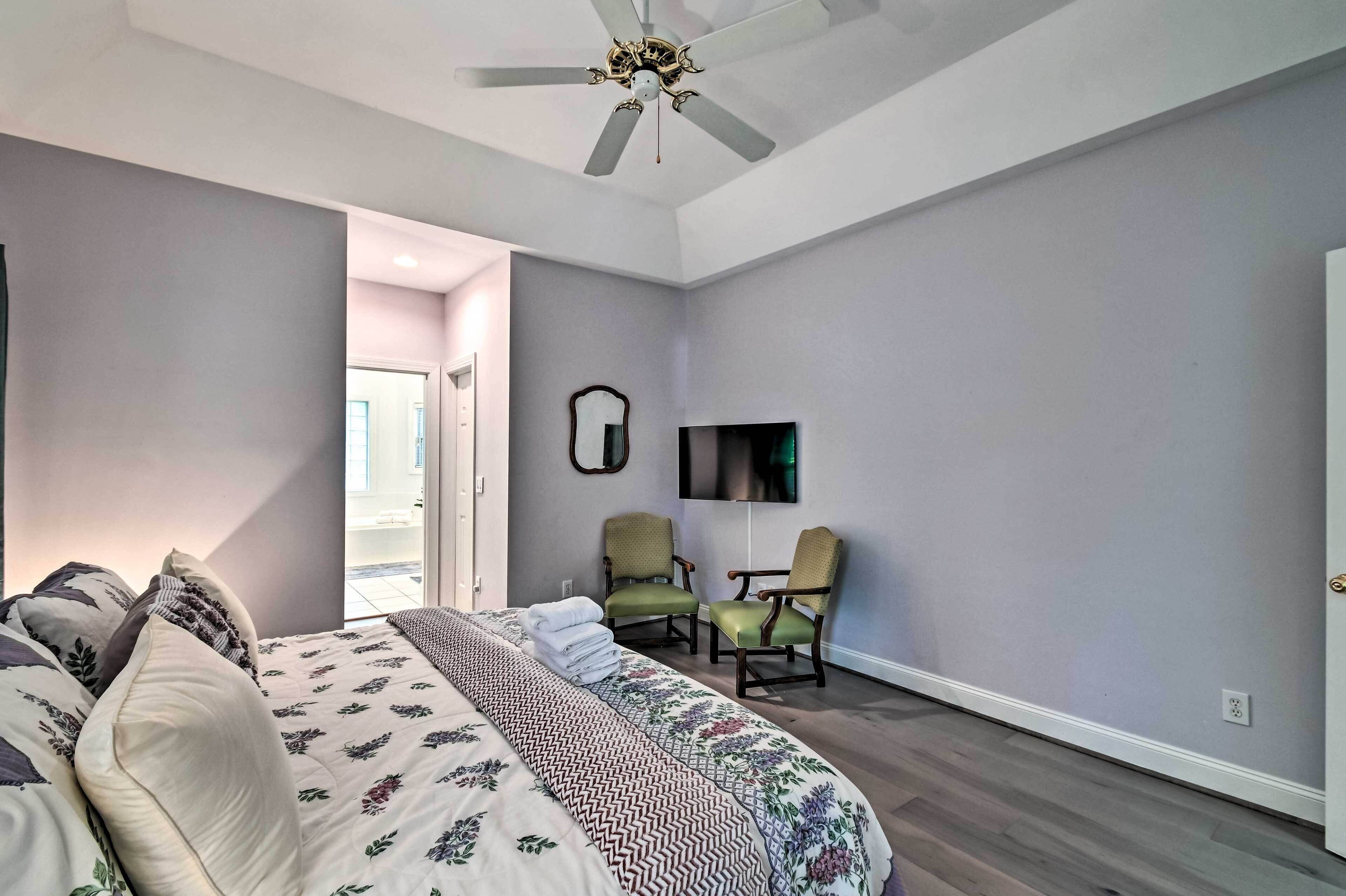 A bedroom with a bed, two chairs, a ceiling fan, and a TV on the wall.