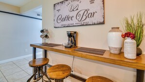 Angolo cottura con cartello che indica una caffetteria, una moka, sgabelli e un vaso con fiori.