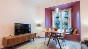 A modern living room with a flat-screen TV, wooden dining table, and a sofa by a large window.