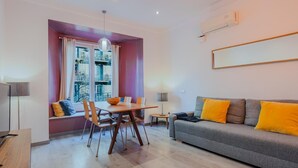 A modern living room with a dining area, a sofa with yellow cushions, and a view of buildings through a window.