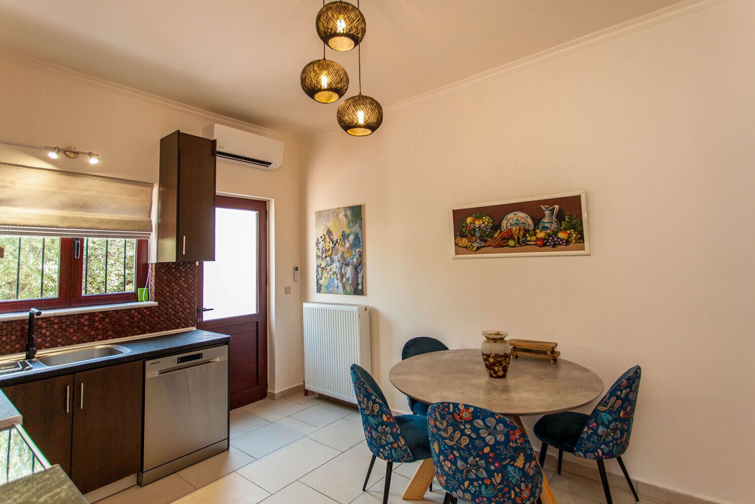 Een moderne keuken met een geïntegreerde vaatwasser, een eethoek met een ronde tafel en gestreepte stoelen, en een schilderij aan de muur.