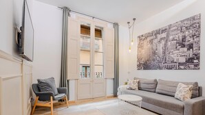 Ein Wohnzimmer mit einer Couch, einem Sessel, einem Couchtisch, einem Fernseher, einem Fenster mit Vorhängen und einem Stadtbild.
