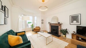 Een ruime woonkamer met een groene bank, een gele fauteuil, een open haard en een televisie.