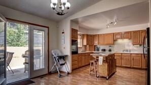 A spacious kitchen with wooden cabinets, a central island, and a sliding glass door leading to an outdoor patio with a grill. - Spacious 5 BDR 3 BA House with Hot Tub, BBQ, Fire Pit, Home Gym, Mountain View (Colorado Springs)