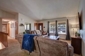 A living room with a brown sofa, a coffee table, and a TV. There is a window with curtains, a door, and a staircase. - Spacious 5 BDR 3 BA House with Hot Tub, BBQ, Fire Pit, Home Gym, Mountain View (Colorado Springs)