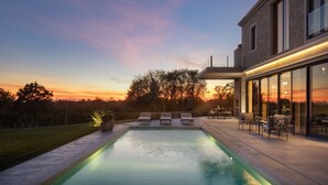 Un área de piscina moderna al aire libre con sillas de descanso y una mesa de patio, rodeada por un jardín ajardinado y un edificio con grandes ventanales.