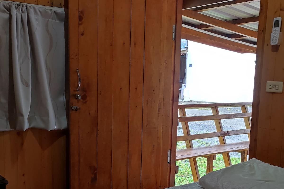 A wooden cabin room with a bed, a wooden door, and a view of a wooden deck outside.