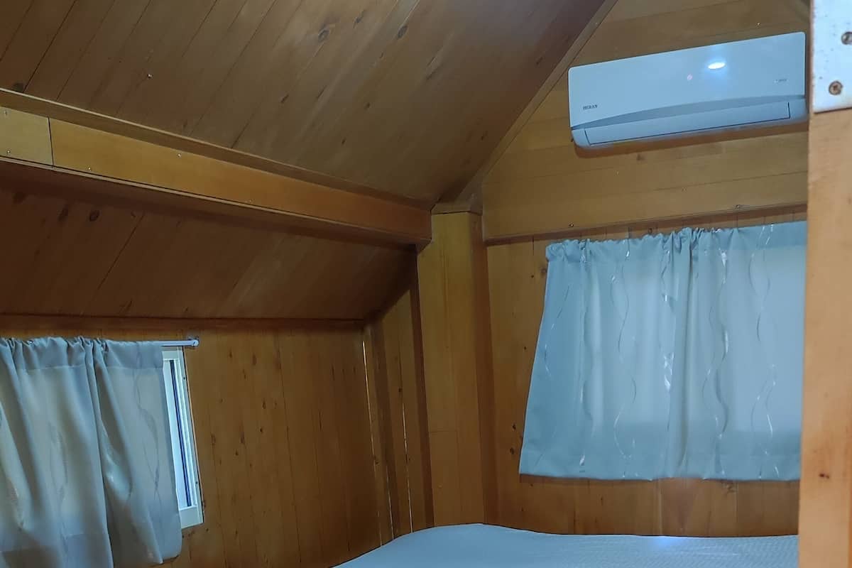 A small, wooden cabin room with a bunk bed, a window with curtains, and a ceiling vent.