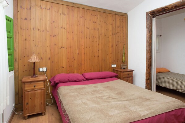 Un dormitorio con pared de madera, una cama con manta color beige, una mesita de noche con lámpara y una pequeña mesa con un florero.