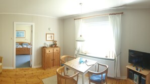 Una habitación acogedora con suelo de madera, cama, una mesita con sillas, televisor y ventana con cortinas.