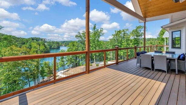 A wooden deck with a view of a lake and forest, a dining area with outdoor furniture, and a covered section with a wooden ceiling. - Coveside Spacious 5BR Waterfront Home (Six Mile)