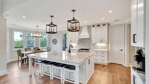 A modern kitchen with white cabinetry, a large island with bar stools, pendant lights, and a view of the outdoors through large windows. - Coveside Spacious 5BR Waterfront Home (Six Mile)