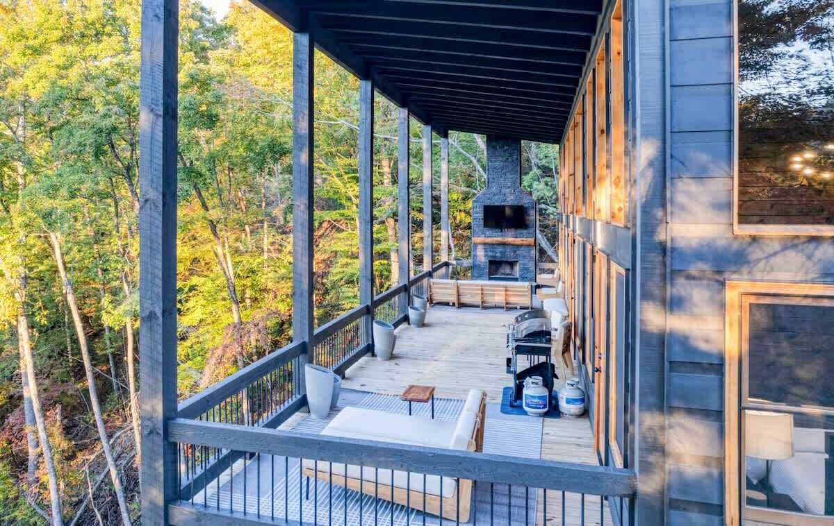 Une terrasse en bois avec des sièges d’extérieur, une cheminée et une vue sur une zone boisée.