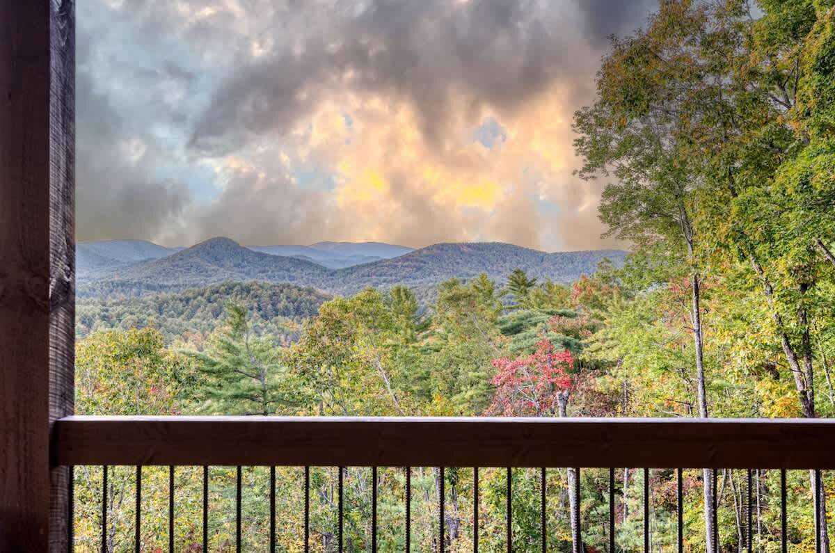 Une vue depuis un balcon doté d’une balustrade, donnant sur un paysage montagneux avec des arbres et un ciel nuageux.