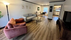 Un salon spacieux avec un canapé gris, un fauteuil rose et une table basse en verre. On y trouve également un coin repas avec une petite table et des chaises, et une cuisine visible à travers une porte ouverte.