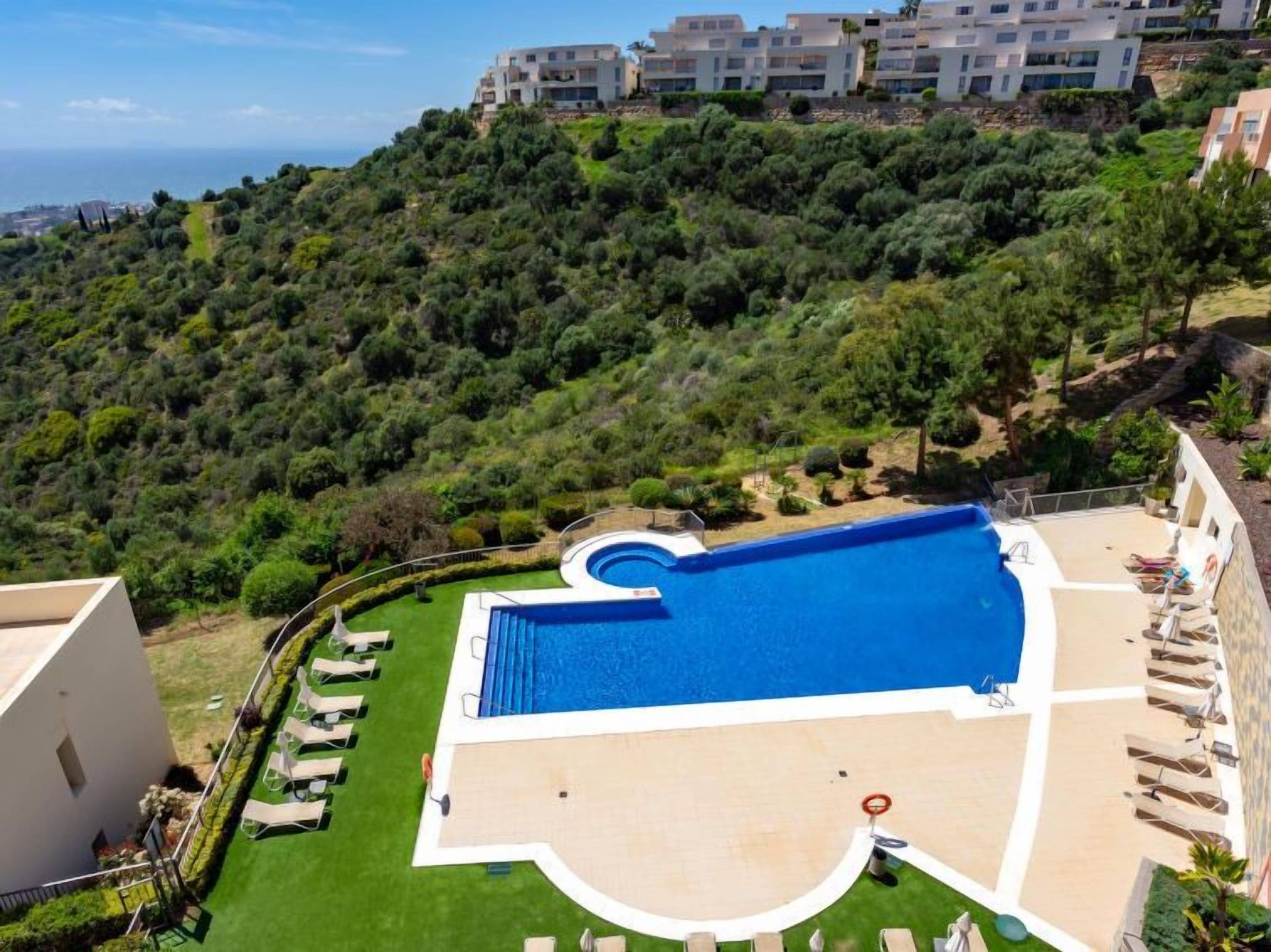 A modern hotel with a large swimming pool, sun loungers, and a view of a lush hillside.