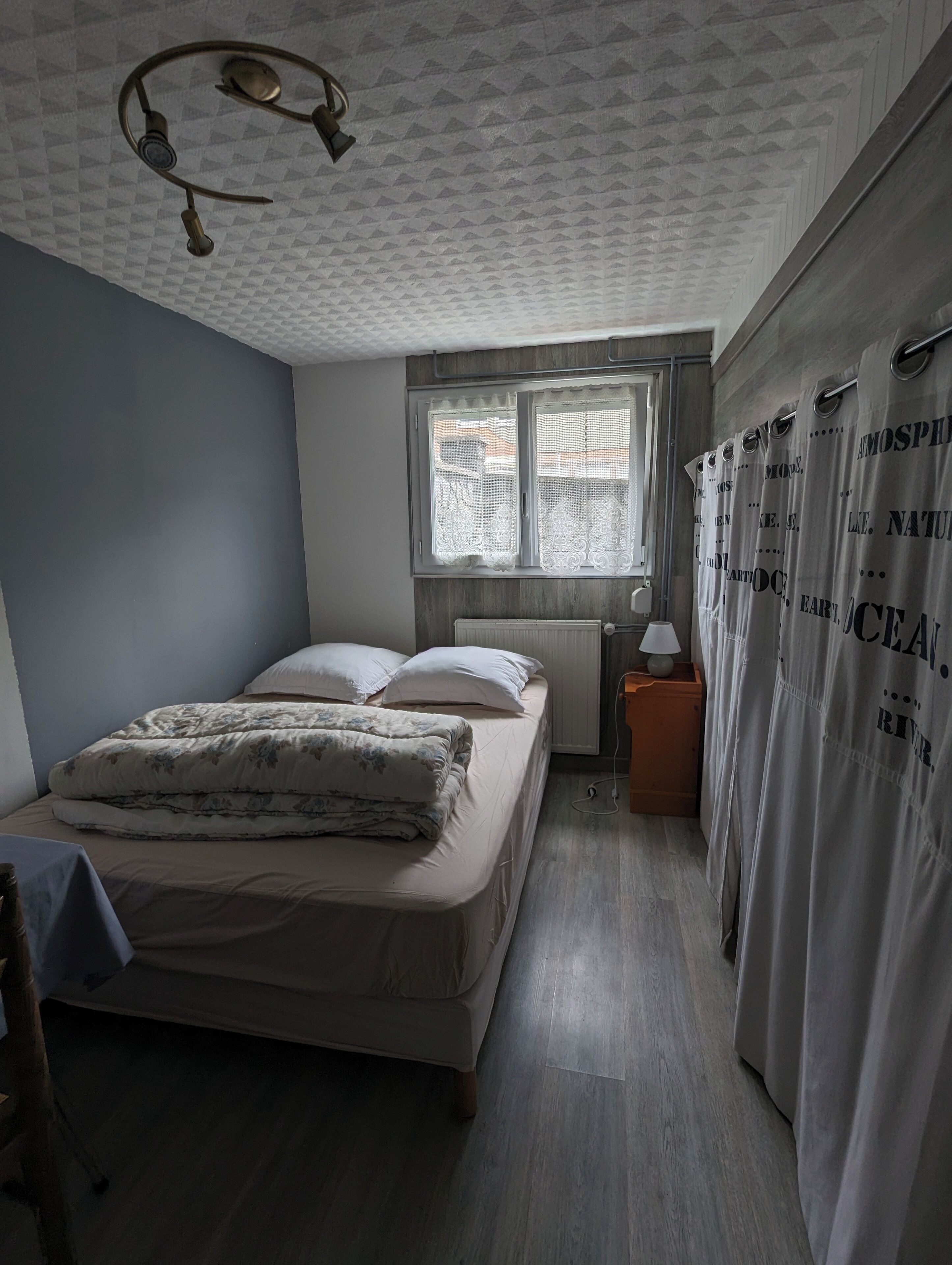 Un dormitorio con cama, ventana, mesita de noche y cortina.