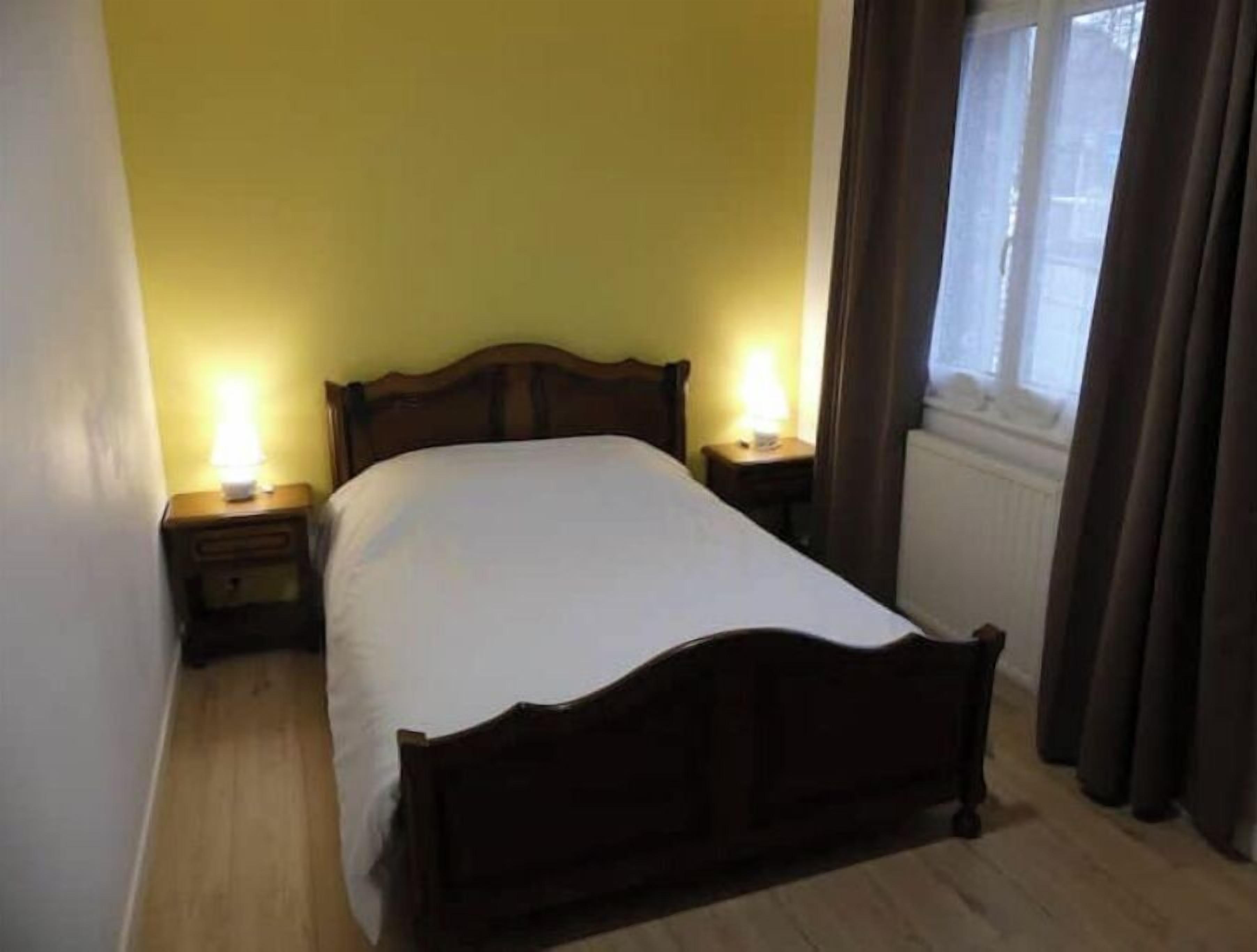 Un lit simple dans une chambre aux murs jaunes, deux tables de chevet avec des lampes et une fenêtre avec des rideaux.