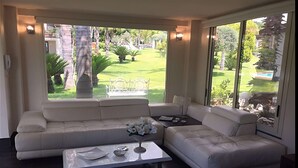 A modern living room with a white sofa and a coffee table, large windows with a view of a garden.