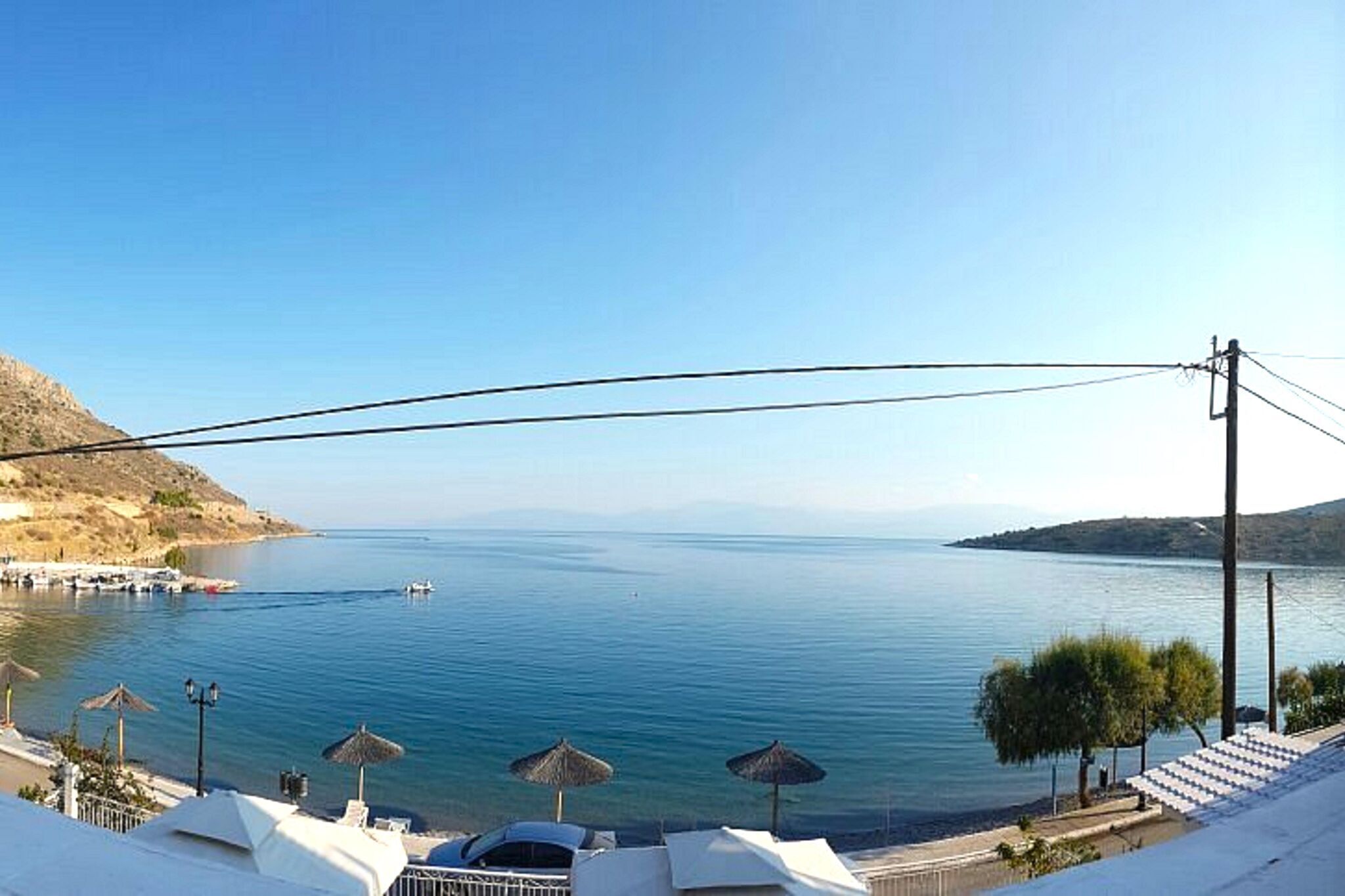 Blick auf die Küste mit blauem Himmel, ruhiger See und vereinzelten Booten. Sonnenschirme und ein weißes Gebäude mit Balkon mit Blick aufs Wasser.