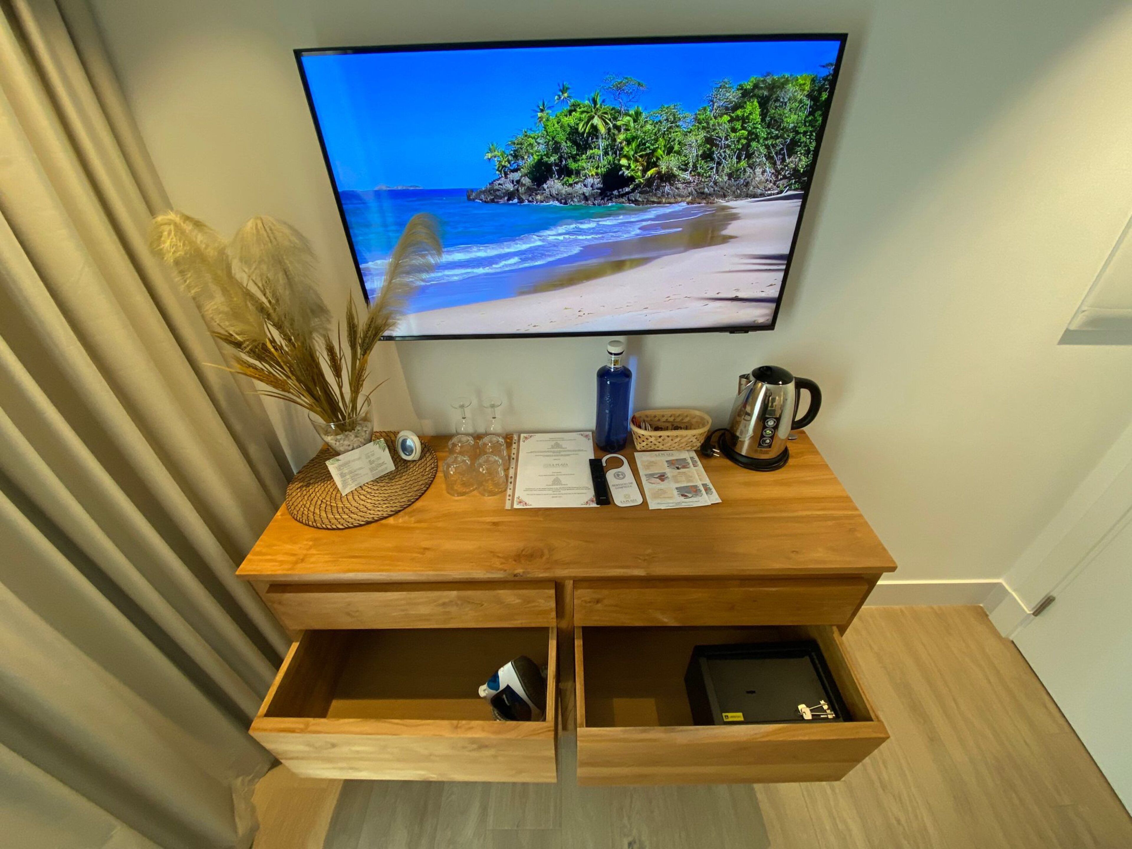 Una consola de madera con un televisor de pantalla plana mostrando una escena de playa, un florero con plantas secas, una cafetera y una cesta.