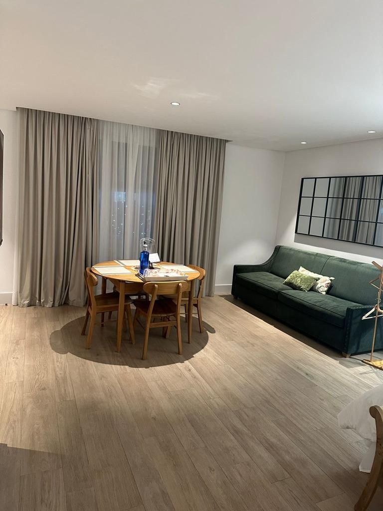 Een moderne woonkamer met een ronde houten tafel, een groene bank en een groot raam met gordijnen.