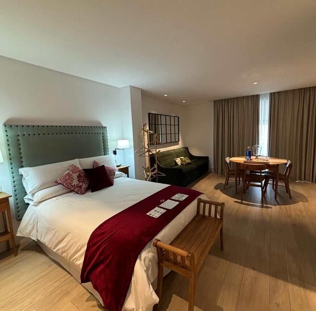 Una habitación de hotel con cama, sofá verde, mesa de comedor con sillas y un banco de madera.