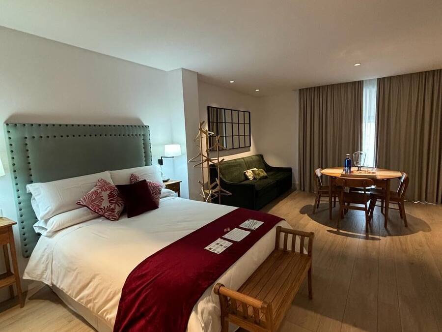 Una habitación de hotel con cama, sofá verde, mesa de comedor con sillas y un banco de madera.