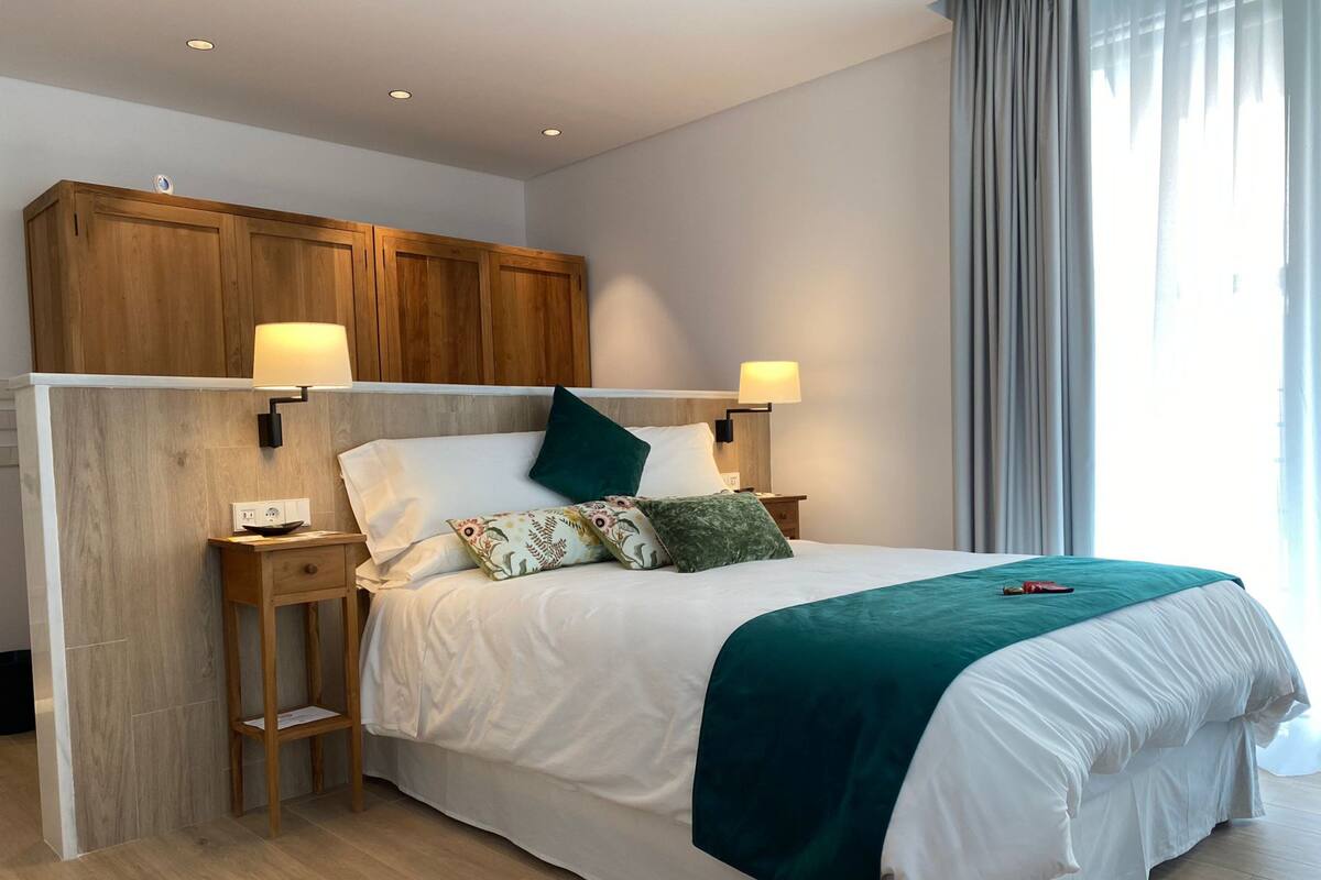 Ein modernes Schlafzimmer mit einem Holz-Kopfteil, zwei Nachttischlampen, einem Bett mit weißer Bettwäsche und einer türkis-farbenen Tagesdecke, sowie einem Nachttisch mit Lampe.