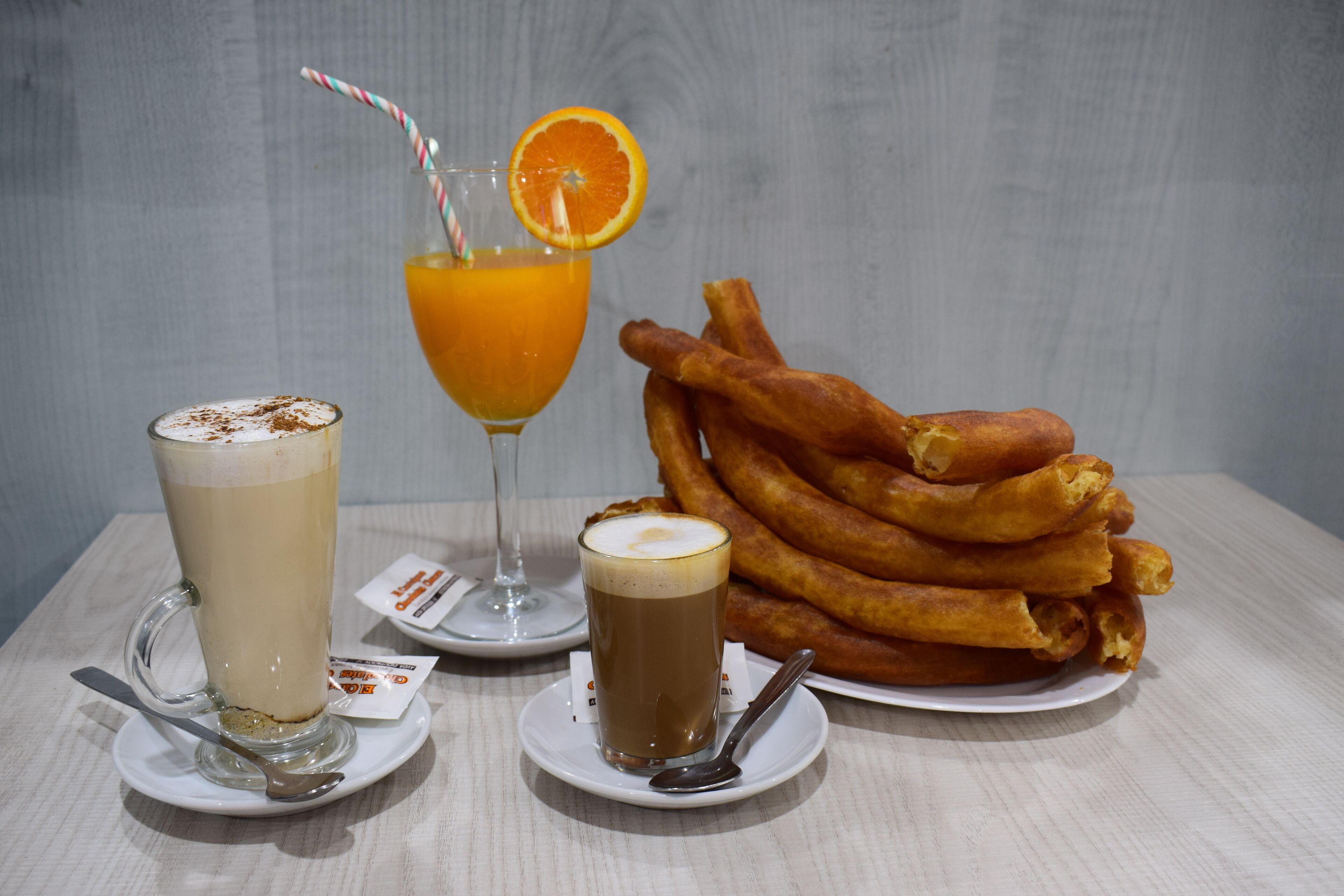 Una mesa con una taza de café, un vaso de jugo de naranja con pajita y un plato de buñuelos.