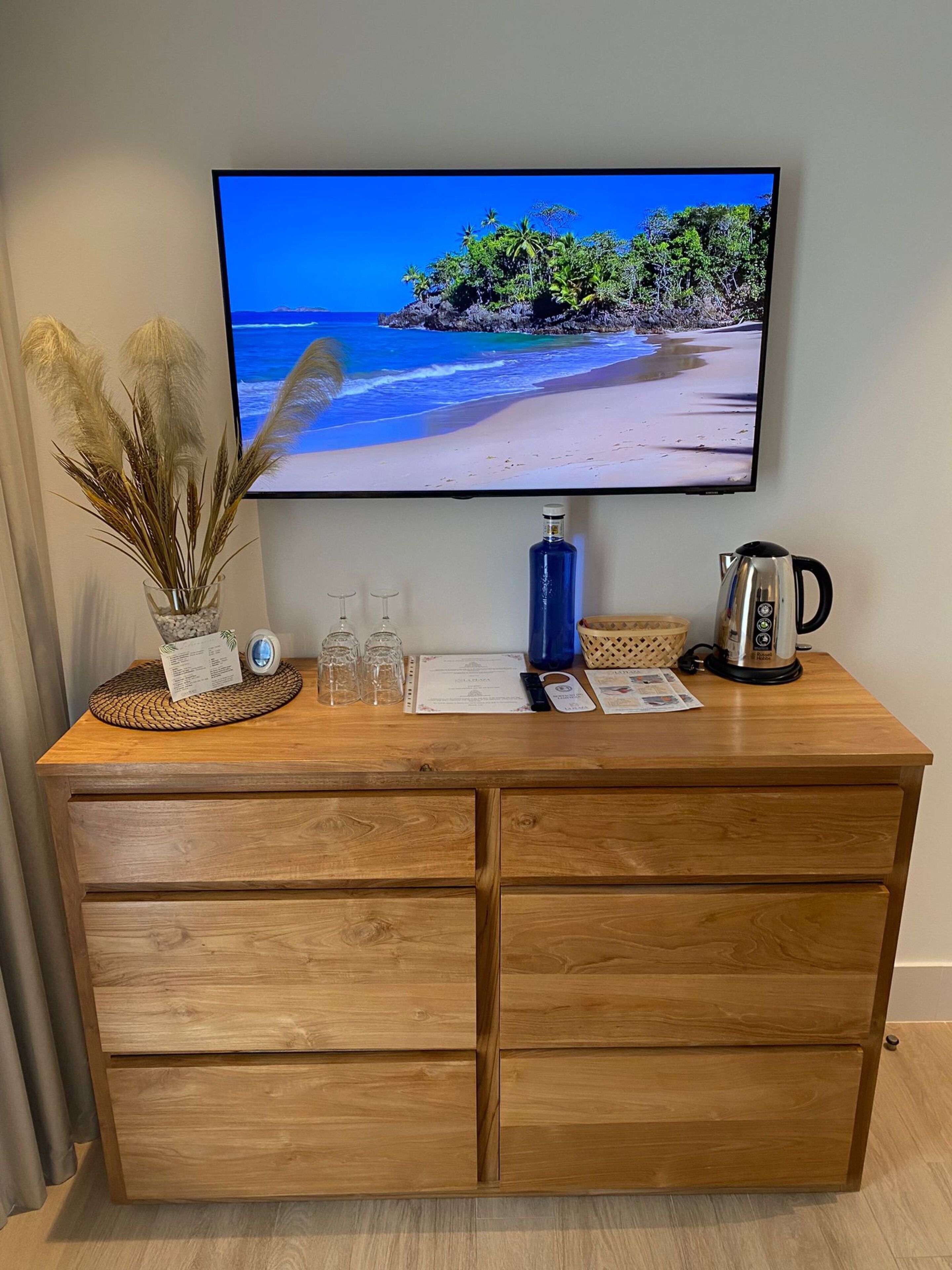 Uma mesa de madeira com uma televisão mostrando uma cena de praia, um vaso com plumas de capim-do-campo e uma cafeteira.