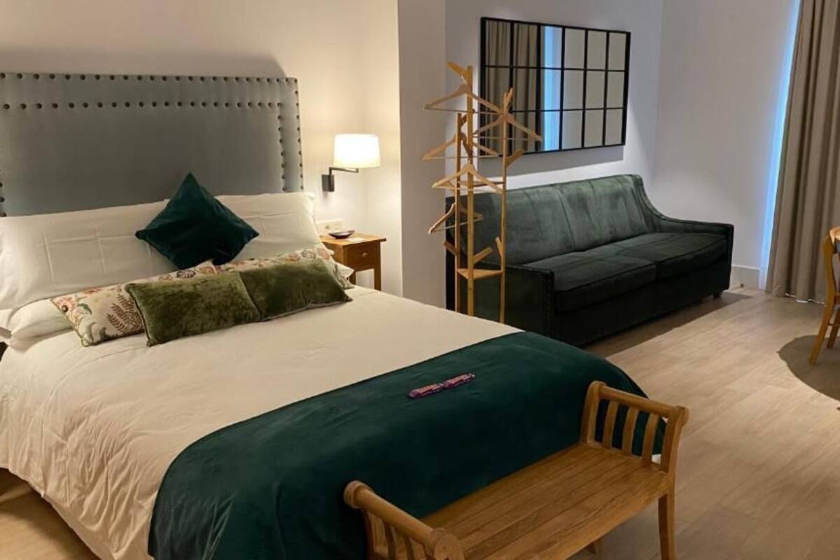 Ein Hotelzimmer mit einem großen Bett, einem grünen Sofa, einem hölzernen Stuhl, einem kleinen Tisch und einem an der Wand befestigten Kofferhaken.