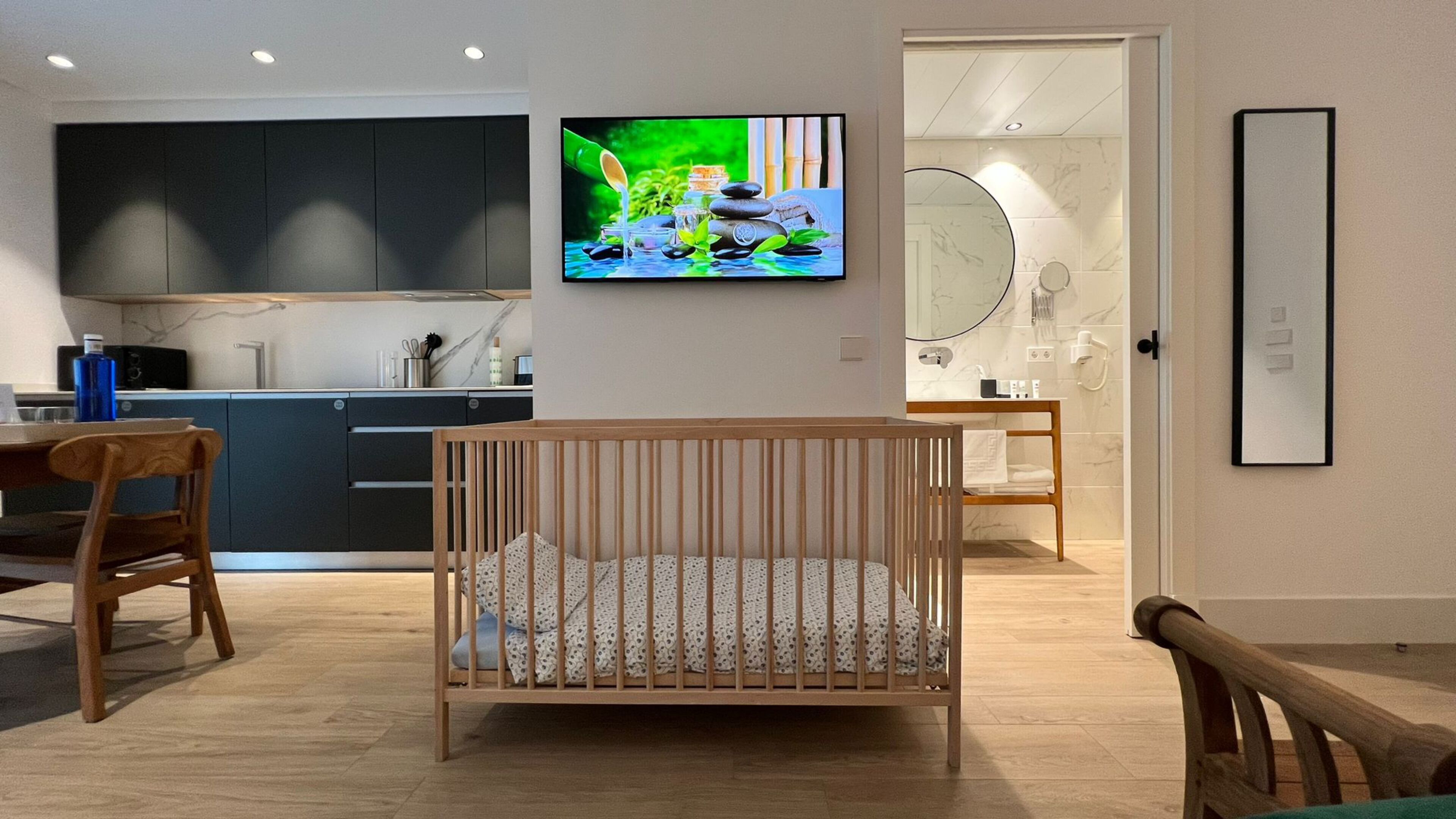 A modern living room with a wooden crib, a dining area with a table and chairs, a kitchen with dark cabinets, and a bathroom with a mirror and towel rack.