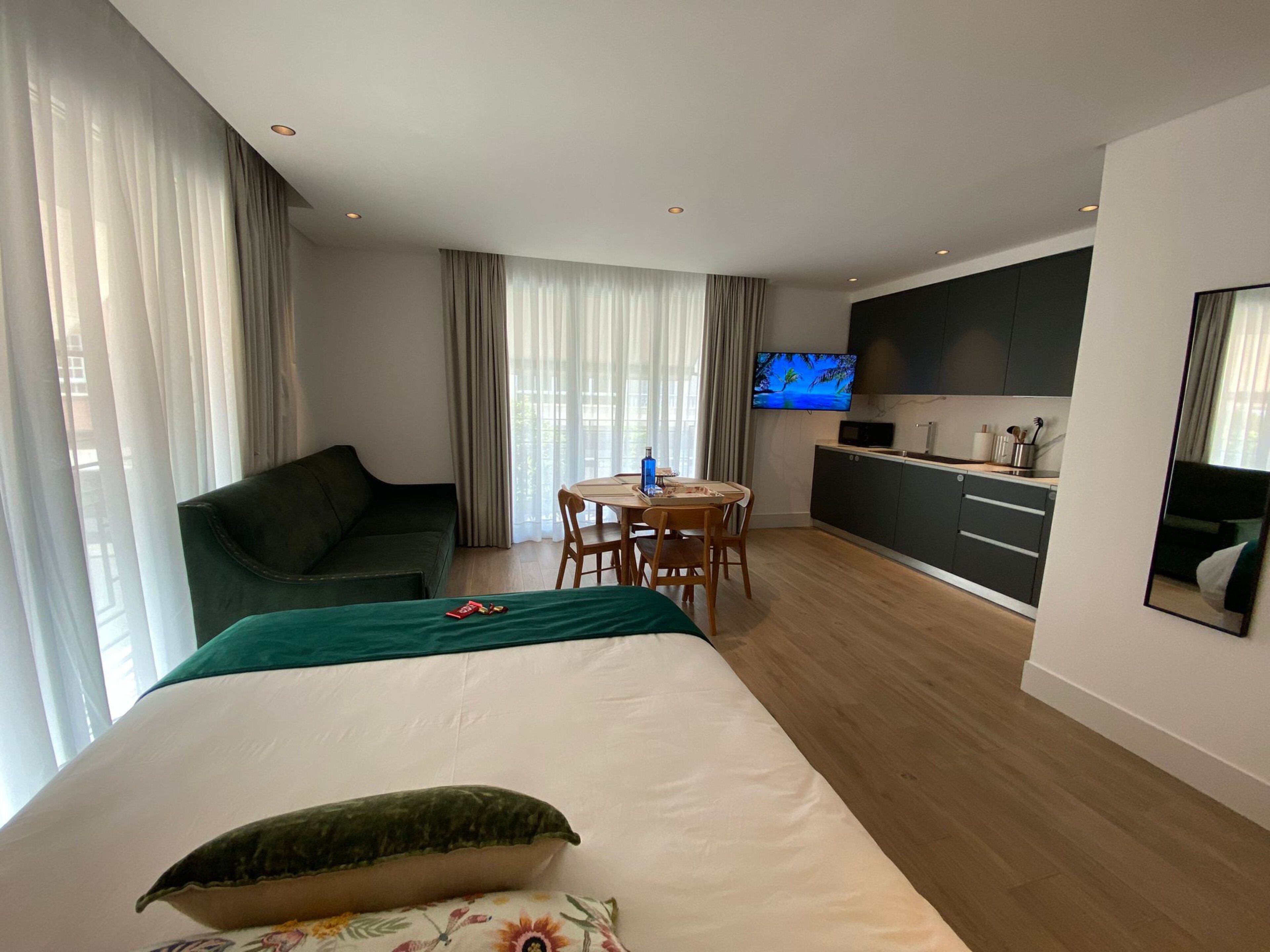 A modern hotel room with a bed, a green sofa, a dining table with chairs, a kitchen area with a TV, and large windows with curtains.