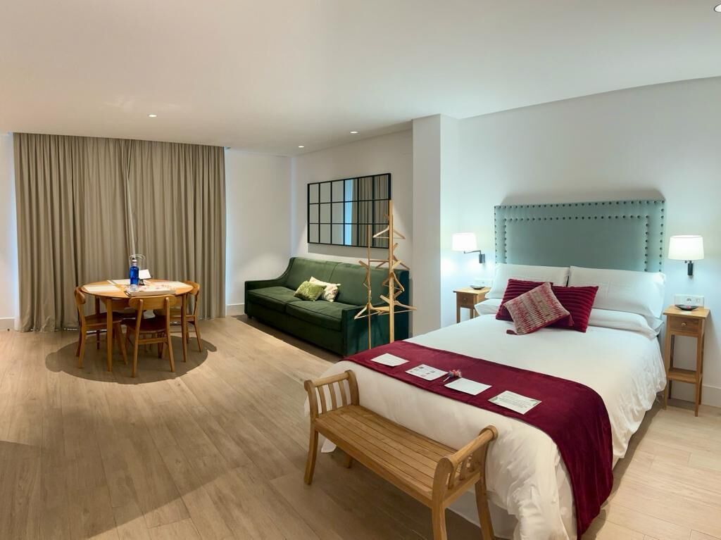 A hotel room with a bed, a green sofa, a round dining table with chairs, a bench, and a small side table with a lamp.
