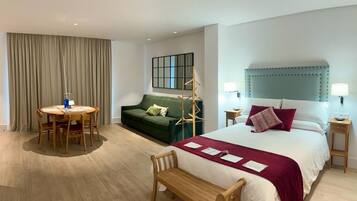 A hotel room with a bed, a green sofa, a round dining table with chairs, a bench, and a small side table with a lamp.