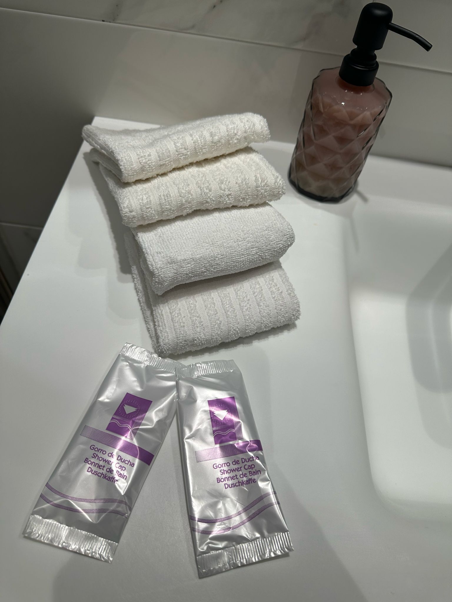 A bathroom with a white sink, a stack of white towels, a bottle of liquid soap, and two packets of shower cap.